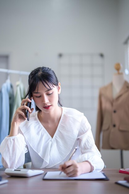 Mujeres comerciantes asiáticas jóvenes que usan su teléfono inteligente para hablar sobre información de clientes y pedidos de ropa para tomar notas mientras trabajan y envían entregas para negocios de compras en línea en la oficina en casa