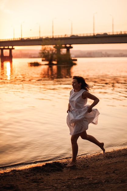 Mujeres corriendo al atardecer una mujer atractiva con un vestido blanco de verano corriendo a lo largo de la orilla del agua en ...