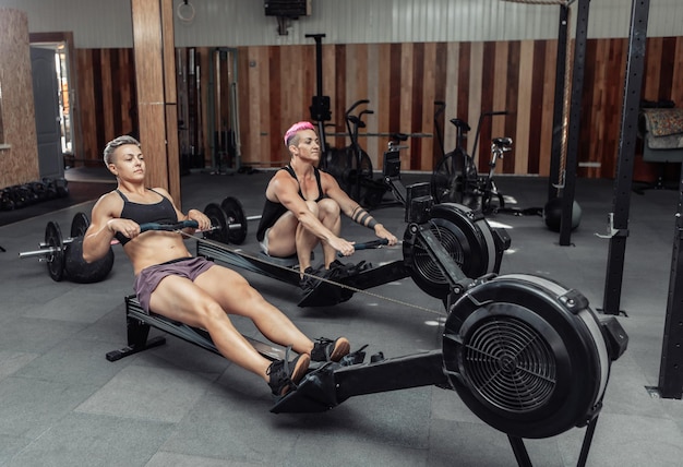 Las mujeres deportivas musculares entrenan en un simulador de entrenamiento en el gimnasio crossfit. Deportistas haciendo ejercicio en máquinas de remo