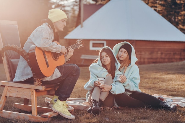 Mujeres encantadoras con sudaderas azules idénticas pasan tiempo en un campamento forestal en el patio del campamento y escuchan la música de un tipo que toca una guitarra acústica junto a ellas.