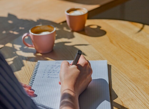 Las mujeres escriben a mano planes en un cuaderno o planificador sobre una mesa de café de madera con luz solar desde la ventana y ...