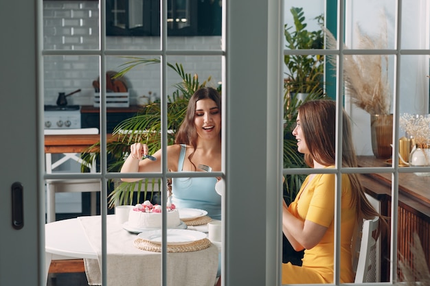 Mujeres felices amigos en casa sentados y sonriendo con pastel de cumpleaños blanco detrás de la puerta de cristal.