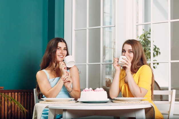 Mujeres felices amigos en casa sentados y sonriendo con pastel de cumpleaños blanco y taza de té.