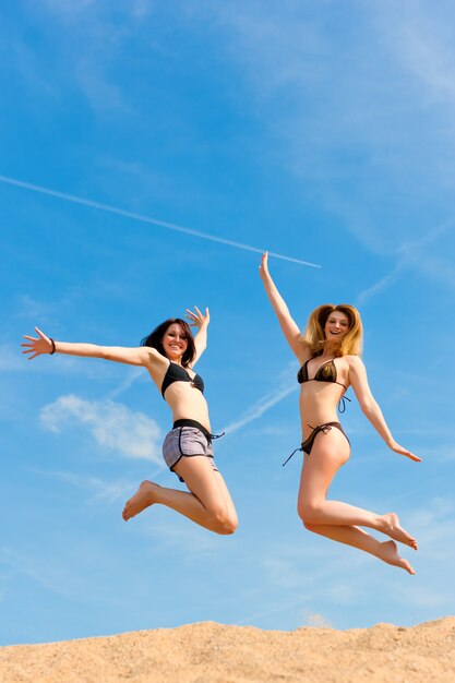 Mujeres felices en traje de baño saltando muy por encima de la arena