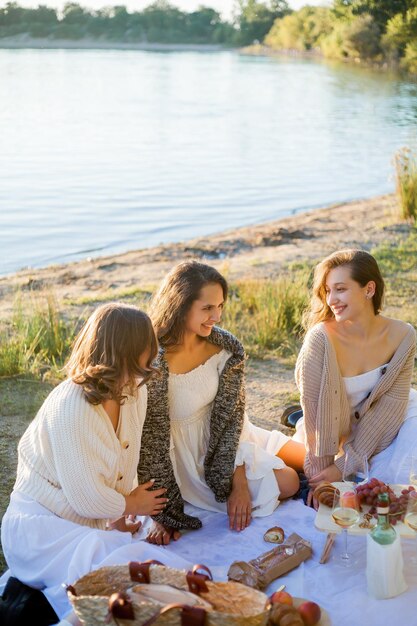 Foto mujeres hermosas jóvenes de 25 años en un picnic de otoño cerca del lago vaso de pasteles de vino blanco