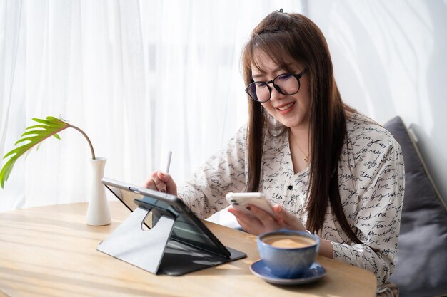 Mujeres independientes asiáticas que escriben un mensaje de negocios en un teléfono inteligente que trabaja tomando notas en una tableta digital con un bolígrafo electrónico para navegar por Internet, chatear y bloguear con una taza de café