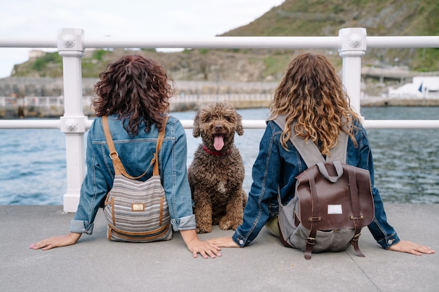 Mujeres irreconocibles con perro de agua marrón. Vista horizontal de mujeres que viajan con mascota. Estilo de vida con animales al aire libre.