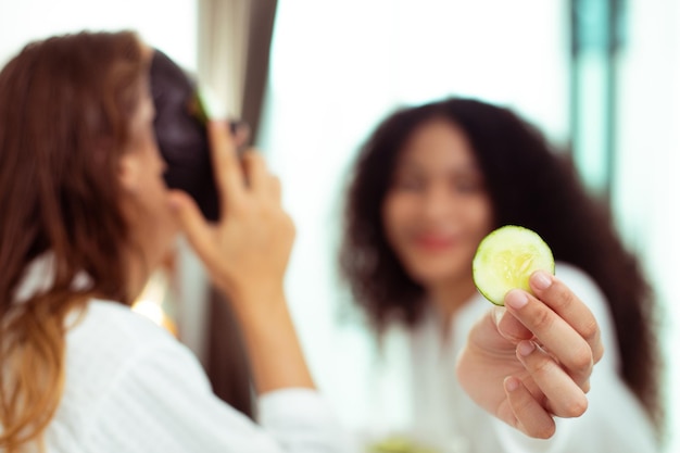 Las mujeres jóvenes adultas caucásicas y afroamericanas aplican una máscara facial de pepino para los ojos y la piel para el tratamiento facial juntos Las parejas LGBT se sienten divertidas y felices con el cuidado de la piel relajándose juntos copiando espacio