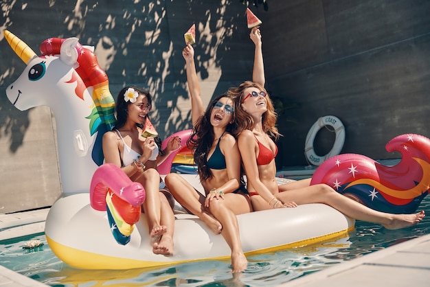 Mujeres jóvenes atractivas en traje de baño sonriendo y comiendo sandía mientras flotan en unicornio inflable en la piscina al aire libre