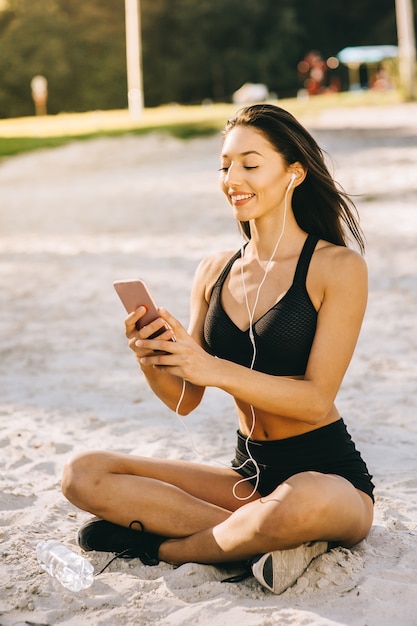 Las mujeres jovenes bastante sonrientes sonrientes del estilo deportivo broncearon vestido en el bikini negro que se sentaba en una playa