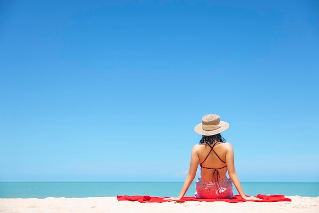 Las mujeres jóvenes en bikini y sombrero de paja se sientan en la playa tropical disfrutando de la vista del océano de la playa