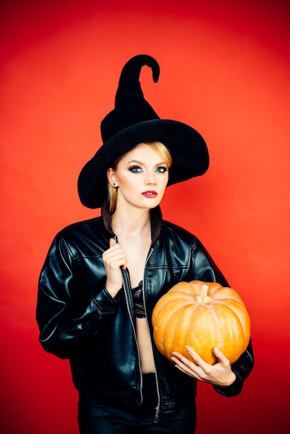 Mujeres jóvenes en disfraces de halloween de bruja negra en fiesta sobre pared roja. Bruja posando con calabaza.