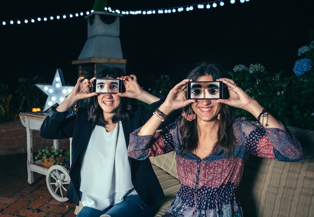 Foto mujeres jóvenes divertidas con teléfonos inteligentes que muestran ojos masculinos en la pantalla en una fiesta al aire libre