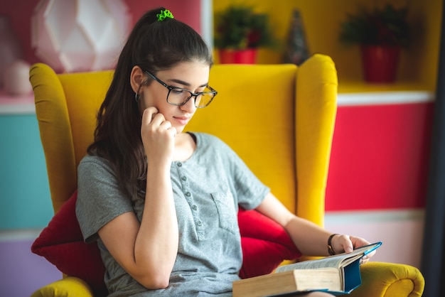 Las mujeres jóvenes están leyendo la literatura de un libro en un sofá amarillo en la sala de estar de la casa, la joven con gafas mientras lee una novela, el concepto de estilo de vida, se relaja y se divierte, el hogar se aísla