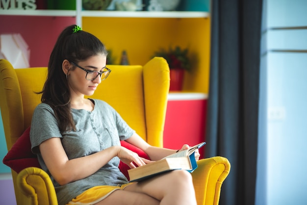 Las mujeres jóvenes están leyendo la literatura de un libro en un sofá amarillo en la sala de estar de la casa, la joven con gafas mientras lee una novela, el concepto de estilo de vida, se relaja y se divierte, el hogar se aísla