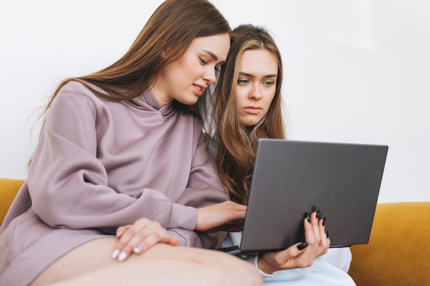 Mujeres jóvenes hermanas gemelas adolescentes con capucha hermosas chicas usando una computadora portátil sentada en un sofá amarillo