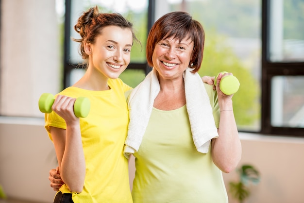 Mujeres jóvenes y mayores en entrenamiento de ropa deportiva con pesas en el interior en el fondo de la ventana