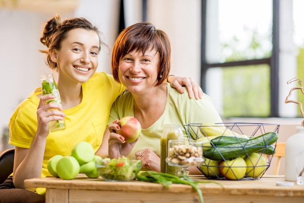 Mujeres jóvenes y mayores sentadas con alimentos saludables y bebidas frescas después del entrenamiento deportivo en el fondo de la ventana