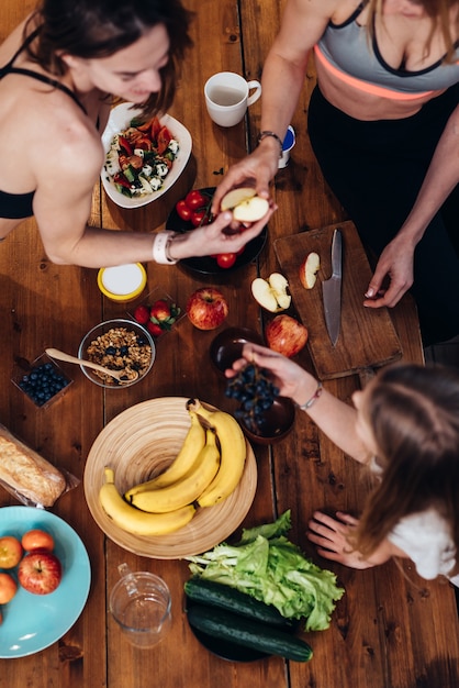 Mujeres jóvenes en ropa deportiva comiendo manzanas, uvas en la cocina.