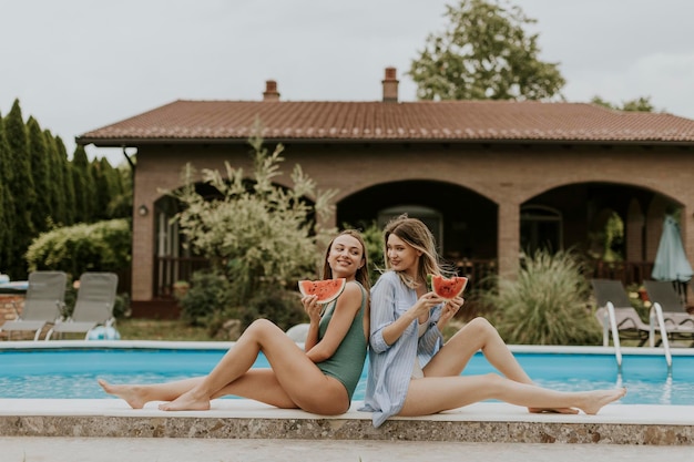 Mujeres jóvenes sentadas junto a la piscina y comiendo sandía en el patio trasero de la casa