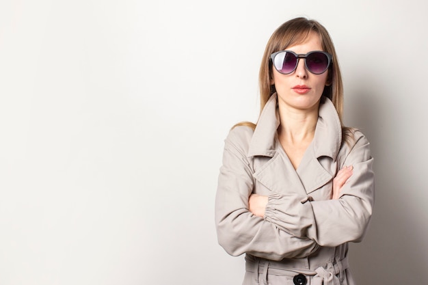 Mujeres jóvenes serias con gafas de sol negras y una capa beige, con los brazos cruzados sobre el pecho. La modelo es obstinada y confiada, posando en una pared luminosa. Espacio vacío para texto