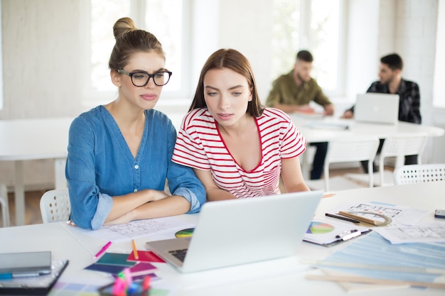 Mujeres jóvenes trabajando cuidadosamente juntas con una computadora portátil mientras pasan tiempo en una oficina moderna