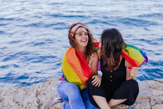 Foto mujeres lesbianas sentadas con una bandera arco iris en la playa