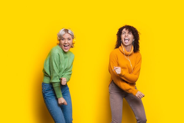 Mujeres locas con auriculares están bailando en una pared de estudio amarillo vistiendo ropa casual y sonriendo a la cámara