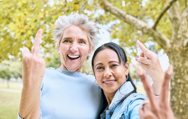 Las mujeres mayores estacionan el retrato y el signo de la mano para un momento divertido y feliz cómico juntos en la naturaleza Ancianas amigas y manos con el dedo medio de la paz y riendo por los árboles, la naturaleza o el bosque para relajarse