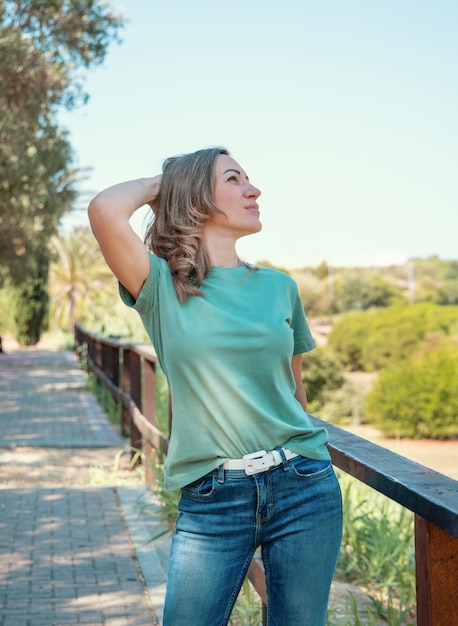 Mujeres de mediana edad vestidas con camiseta y jeans permanece al aire libre en el parque con las manos sobre la cabeza