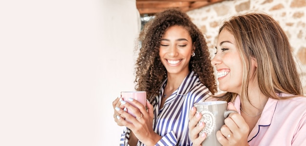 Mujeres multiétnicas mejores amigas en pijama sonriendo sosteniendo una taza de té con espacio de copia en blanco a la izquierda - Concepto: quedarse en casa y disfrutar de su vida