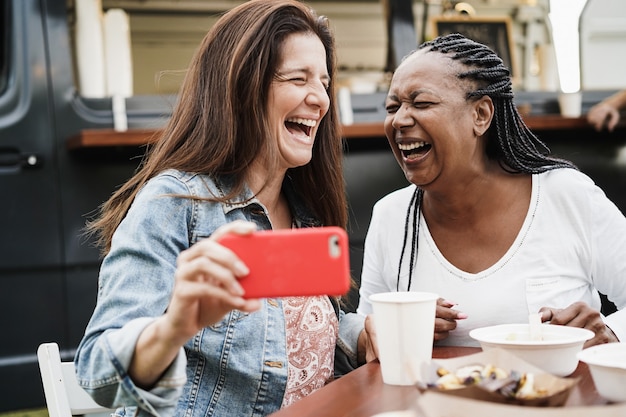 Mujeres multirraciales que se divierten tomando fotos con teléfonos móviles en el restaurante de camiones de comida al aire libre - Concepto de verano y amistad - Enfoque en el rostro femenino afroamericano