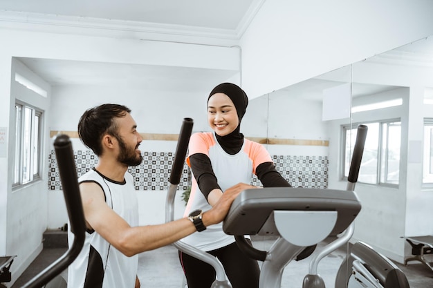 Mujeres musulmanas en el gimnasio haciendo ejercicios cardiovasculares en una máquina de ciclo elíptico estático con ayuda de un amigo h