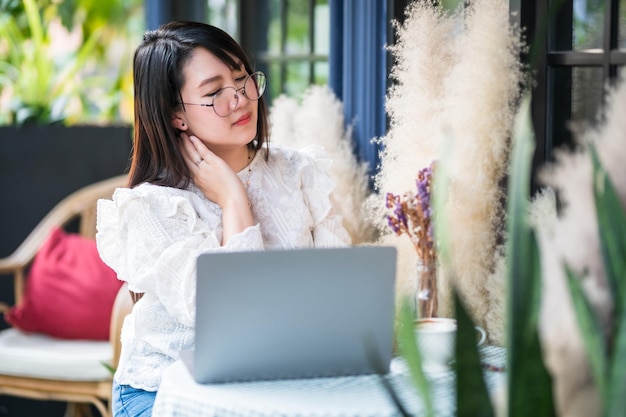 Mujeres de negocios asiáticos independientes tocar masajear el cuello rígido después de trabajar con una computadora portátil con una taza de café y un teléfono inteligente se sintió como relajarse en una cafetería como el fondo