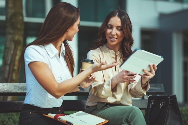 Las mujeres de negocios están sentadas en el parque de oficinas mientras usan una tableta digital y sostienen una taza de café desechable. El equipo de negocios está trabajando juntos en línea mientras consulta.