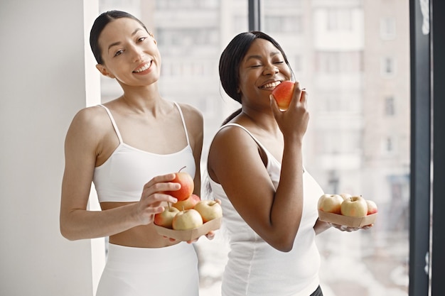 Mujeres negras y caucásicas de pie en un estudio con grandes ventanas y sosteniendo manzanas