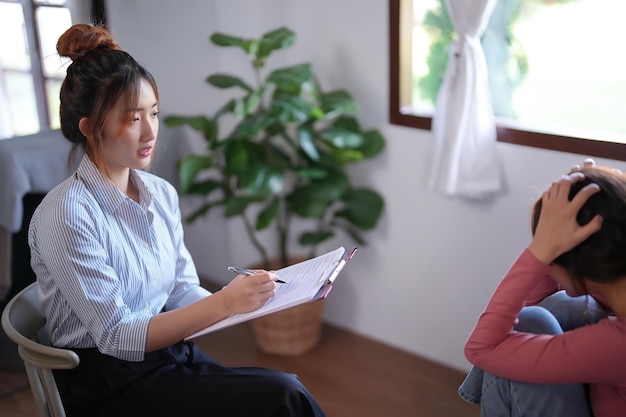 Mujeres pacientes asiáticas estresadas y con dolor de cabeza por ansiedad mientras la psicóloga examina problemas de salud psicológica y escribe notas en papel para asesoramiento sobre terapia de salud mental