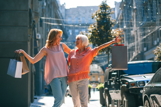 Mujeres positivas de dos generaciones sonriendo el uno al otro y llevando bolsas de la compra.