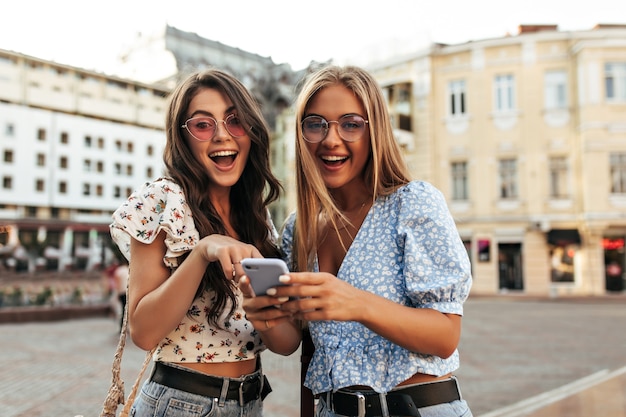 Las mujeres rubias y morenas sorprendidas en blusas florales recortadas de moda de verano y gafas de sol coloridas sonríen ampliamente y sostienen el teléfono púrpura al aire libre