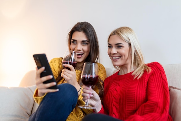 Mujeres sentadas en el sofá riendo en un acogedor loft con vino Dos amigas relajándose en el sofá en casa con una copa de vino hablando juntas