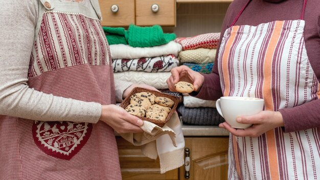 Mujeres sosteniendo una taza de café y galletas vestidas con delantales festivos con un montón de jerseys diferentes en la cocina.