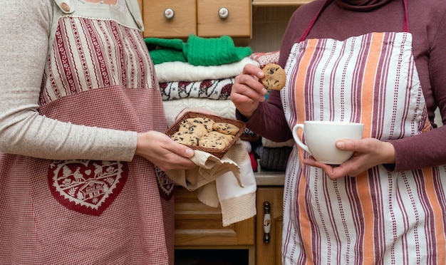 Mujeres sosteniendo una taza de café y galletas vestidas con delantales festivos con un montón de jerseys diferentes en la cocina.