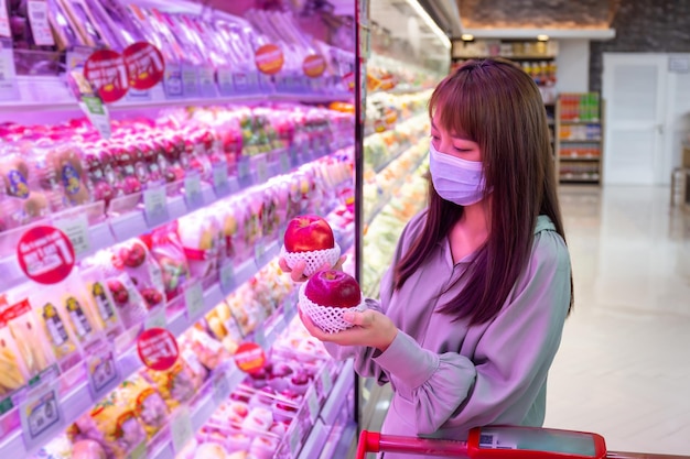 Las mujeres usan máscaras para comprar manzanas en los supermercados, nuevos estilos de vida normales en la era del virus Corona o la epidemia de Covid-19