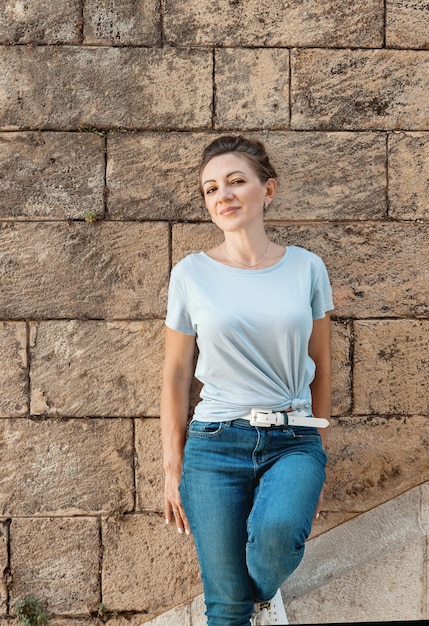 Las mujeres vestidas con camiseta y jeans permanece cerca de una pared al aire libre en la calle