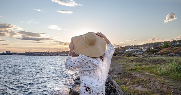 Mulher à beira-mar na vista traseira do pôr do sol. Vestido com uma saia longa e um chapéu.