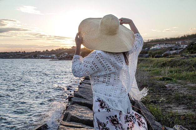 Mulher à beira-mar na vista traseira do pôr do sol. Vestido com uma saia longa e um chapéu.