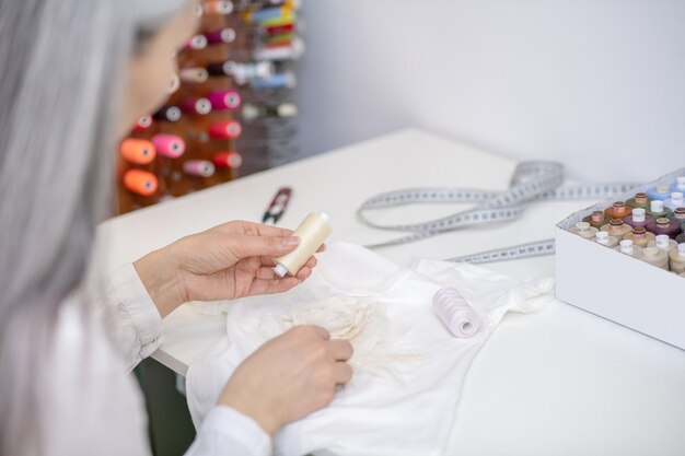 Mulher à mesa segurando um novelo de linha bege sobre a costura, comparando a combinação de cores