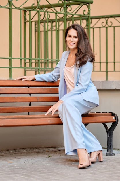 Mulher à moda no pantsuit skyblue que tem o descanso em um banco perto do escritório