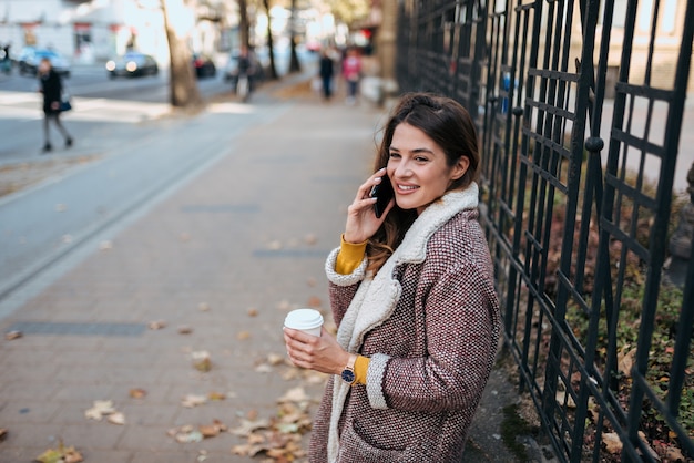 Mulher à moda que fala no telefone e que bebe o café na rua da cidade.