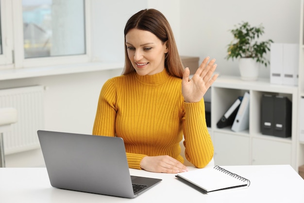 Mulher acenando para cumprimentar durante uma videoconferência via laptop na mesa do escritório